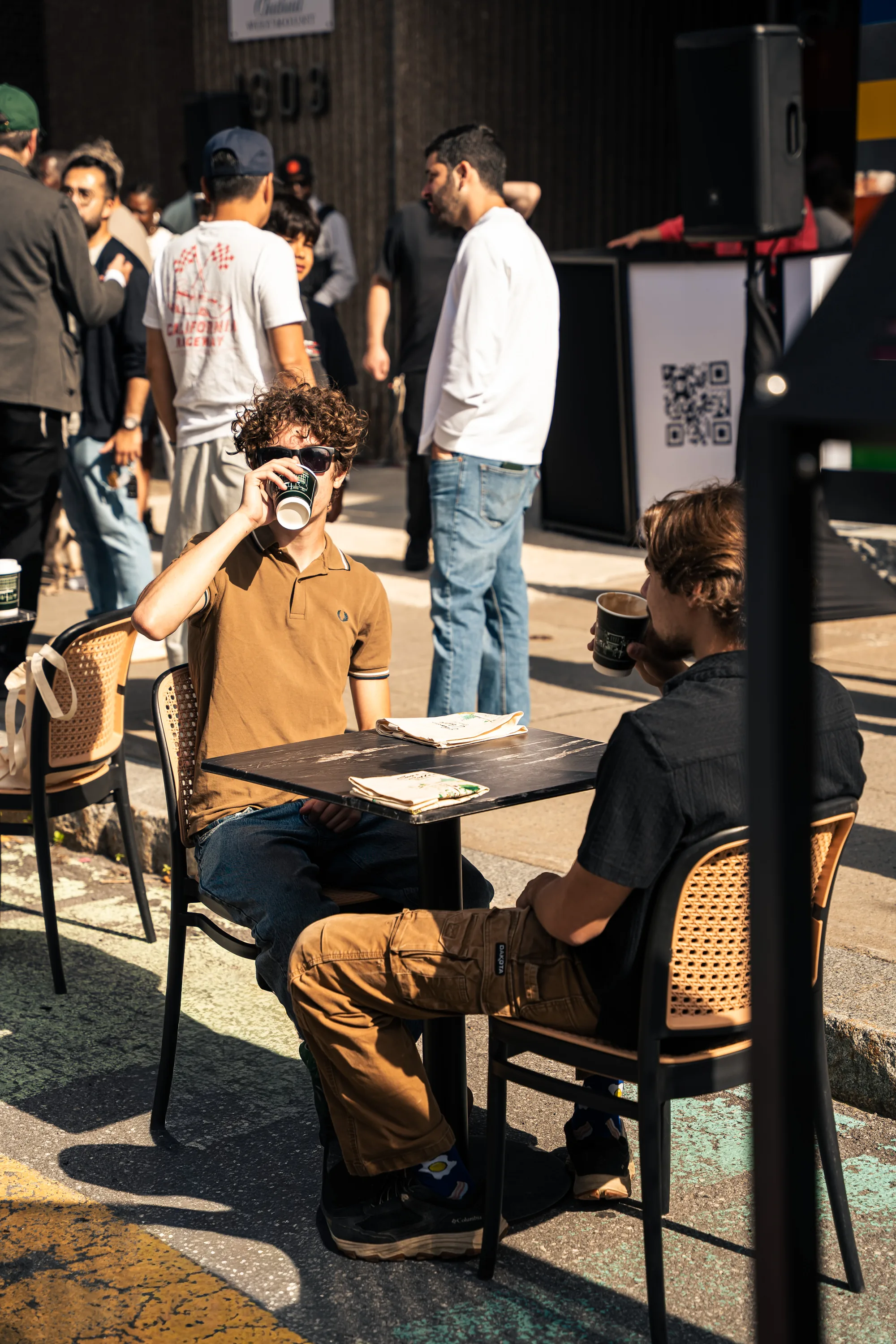 Two men with coffee at a sidewalk table while the crowd stands around them.
